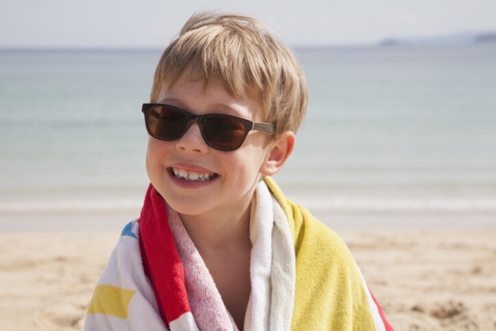 A boy in sunglasses on the beach, with a towel around his shoulders